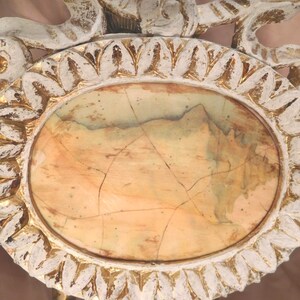 Puede incluir: Una bandeja decorativa ovalada con una incrustaci&oacute;n central de piedra pulida que presenta vetas y grietas naturales en tonos bronceado, beige y verde p&aacute;lido. El marco grueso y ornamentado tiene un acabado de pintura blanca desgastada sobre oro.