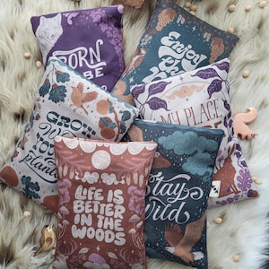 Könnte beinhalten: Eine Sammlung dekorativer Kissen mit von der Natur inspirierten Designs und Texten. Die Kissen zeigen Illustrationen von Vögeln, Hirschen und Pflanzen mit Sätzen wie "Born to be", "Enjoy the" und "Life is better in the woods". Sie sind auf einer weißen Pelzoberfläche angeordnet.