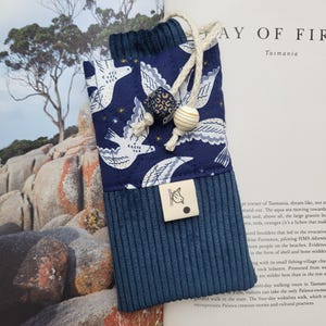 Könnte beinhalten: Ein marineblaues Brillenetui mit Corduroy-Boden und einem gemusterten Oberteil mit weißen Vögeln. Das Etui hat einen Kordelzugverschluss mit dekorativen Holzperlen und ein kleines Etikett mit einer Vogelillustration.