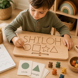 May include: A light-colored wooden tracing board featuring the name SHILOH engraved at the top, followed by engraved shapes including a circle, square, star, and triangle, accompanied by a wooden stylus for tracing and educational cards.