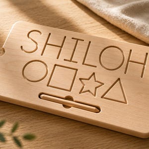 May include: A rectangular light-colored wooden tracing board featuring the engraved name SHILOH at the top. Below the name are four engraved shapes: a circle, a square, a star, and a triangle. A wooden stylus is stored in a horizontal slot.