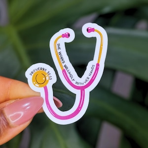 Könnte beinhalten: Ein Aufkleber in Form eines Stethoskops mit rosa und gelben Akzenten. Der Aufkleber trägt den Text "SHE WORKS WILLINGLY WITH HER HANDS" und "PROVERBS 31:13". Das Stethoskop hat rosa Ohrstücke und einen gelben Schlauch.