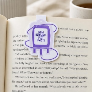 May include: A purple and white bookmark shaped like an IV bag with a tube wrapped around it. The text on the bookmark reads: NOW INFUSING: PLOT TWISTS. The bookmark is placed inside an open book with visible printed text on the pages.