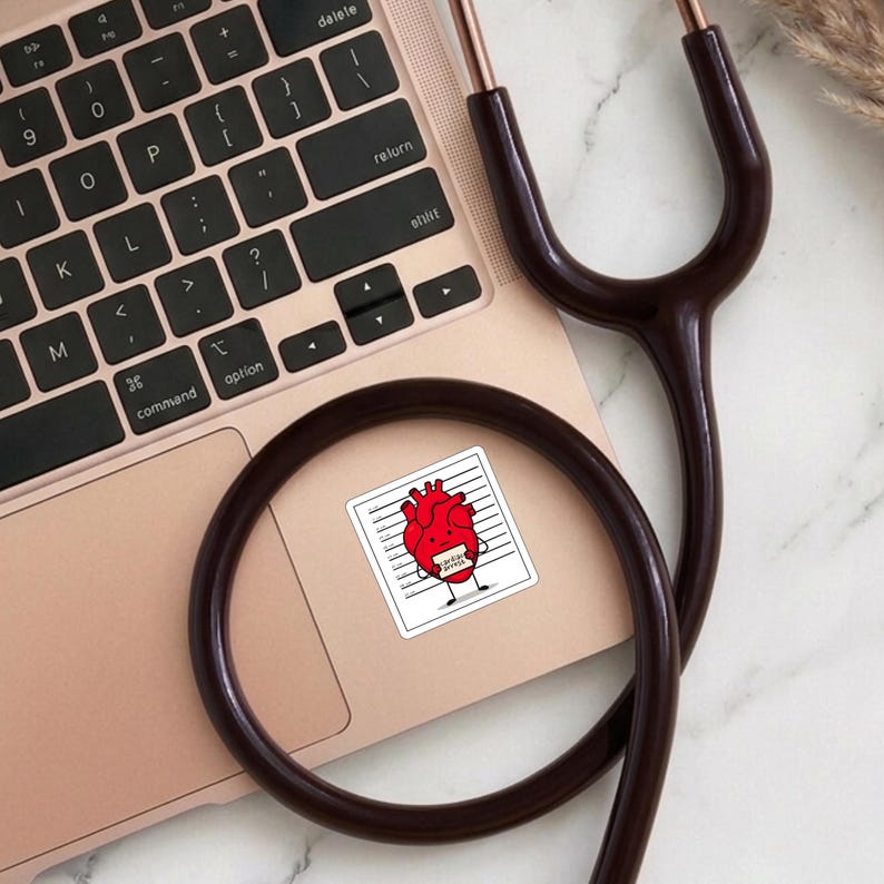 Peut inclure: Un autocollant rouge en forme de c&oelig;ur avec des bras et des jambes, ressemblant &agrave; un personnage de dessin anim&eacute;, est plac&eacute; sur un ordinateur portable. L'autocollant est dans le style d'une ligne de police, avec le texte "Cardiac Arrest". Un st&eacute;thoscope marron est pos&eacute; sur l'ordinateur portable.