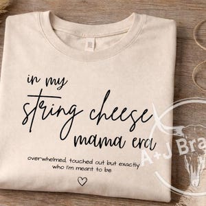 May include: A cream-coloured t-shirt with black text. The text reads "in my string cheese mama era", with the words "overwhelmed touched out but exactly who I'm meant to be" below, and a heart symbol.