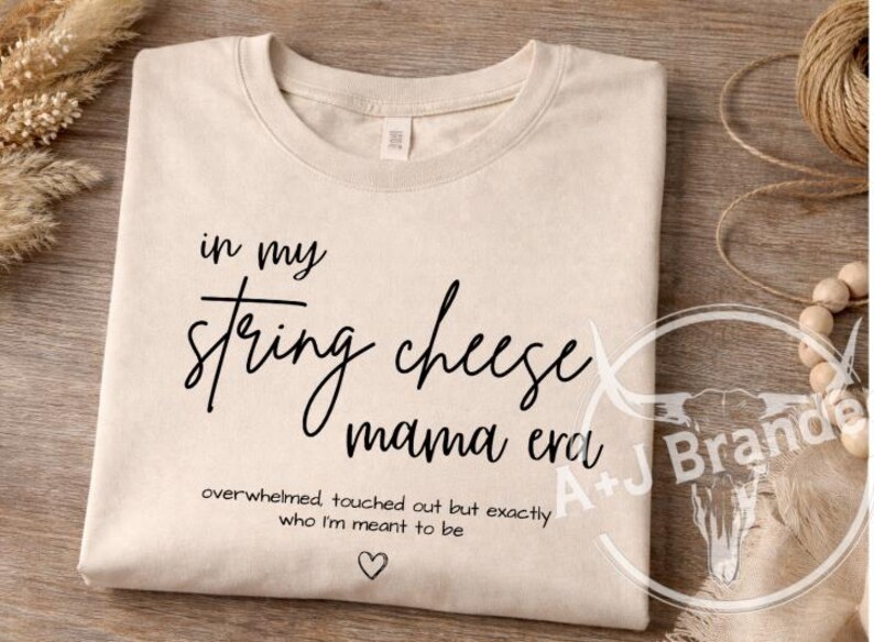 May include: A cream-coloured t-shirt with black text. The text reads "in my string cheese mama era", with the words "overwhelmed touched out but exactly who I'm meant to be" below, and a heart symbol.