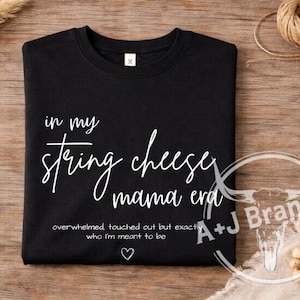 May include: Black crew neck t-shirt with white text that reads "in my string cheese mama era." Additional text below says "overwhelmed touched out but exactly who I'm meant to be." A heart symbol is below the text.