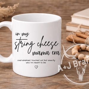 May include: White ceramic mug with the text "in my string cheese mama era" in black script. Additional text below reads "overwhelmed touched out but exactly who I'm meant to be" with a heart symbol. The mug has a curved handle.