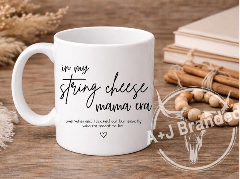 May include: White ceramic mug with the text "in my string cheese mama era" in black script. Additional text below reads "overwhelmed touched out but exactly who I'm meant to be" with a heart symbol. The mug has a curved handle.