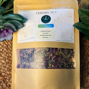 May include: A brown paper pouch containing a Somatic Tea Bath Blend labeled Embody Tea Mermaid Ritual. The label lists ingredients including calendula flowers, rose petals, stinging nettle leaf, and rosemary. A clear window reveals the dried herbs.