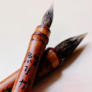 May include: Two calligraphy brushes with natural bamboo handles and dark bristles. The front handle features black Japanese characters engraved into the wood. The brushes have a polished finish and are positioned diagonally against a light background.