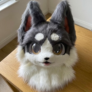 Puede incluir: Cabeza de fursuit de husky de piel sintética gris, blanca y negra. Tiene ojos grandes y expresivos y detalles rosas en el interior de las orejas. El diseño incluye un hocico blanco y una pequeña lengua rosa.