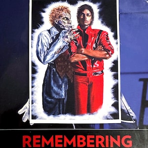 Puede incluir: Impresión con borde blanco que muestra dos figuras: una con un atuendo rojo y la otra como un zombi. El texto "REMEMBERING MICHAEL" está en la parte inferior. La imagen está sobre un fondo azul.