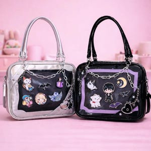 May include: Two square-shaped handbags. One is silver with a silver handle and the other is black with a black handle. Both have a clear front panel with decorative pins and chain accents. The bags are decorated with cartoon-style pins.