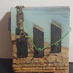 May include: A rustic, handmade wooden art piece. The artwork features a weathered, textured wooden base with a blue and light blue painted background. Two dark rectangular shapes are affixed to the wood, connected by a green wire. A metal clasp is attached to the top.