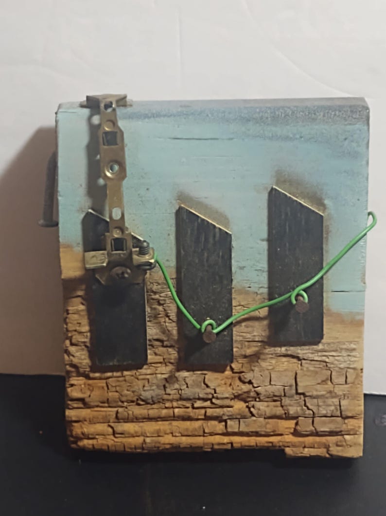 May include: A rustic, handmade wooden art piece. The artwork features a weathered, textured wooden base with a blue and light blue painted background. Two dark rectangular shapes are affixed to the wood, connected by a green wire. A metal clasp is attached to the top.