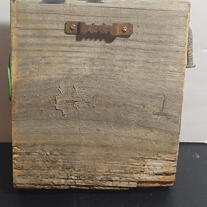 May include: A weathered wooden block with a metal sawtooth hanger on the back. The wood has a gray and brown color with visible grain and cracks. A small metal piece is attached to the top right corner, and a green wire is visible on the left side.