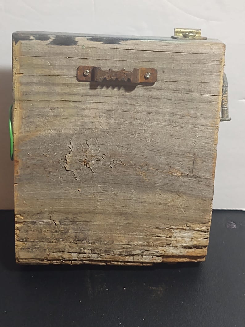 May include: A weathered wooden block with a metal sawtooth hanger on the back. The wood has a gray and brown color with visible grain and cracks. A small metal piece is attached to the top right corner, and a green wire is visible on the left side.