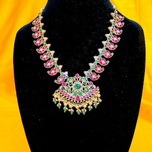 May include: A gold-toned necklace with a central pendant and a series of paisley-shaped links. The necklace features red, green, and white gemstones, with small pearls and green beads. The necklace is displayed on a black mannequin.