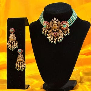 May include: An ornate gold-toned necklace and matching earrings set. The necklace features a central pendant with a deity figure, red and green accents, and dangling pearls. The earrings mirror the pendant design. The jewelry is displayed on black stands against a yellow background.