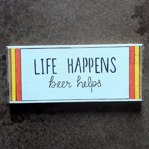 May include: A rectangular wooden sign with the text "LIFE HAPPENS beer helps" in black lettering. The sign has a light blue background and is framed with vertical stripes of red, yellow, and orange.