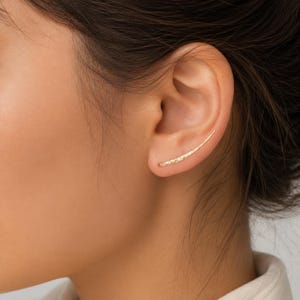 Może przedstawiać: Złoty kolczyk typu ear climber o smukłym, zakrzywionym kształcie, który dopasowuje się do naturalnej linii płatka ucha. Metalowa powierzchnia posiada subtelną, fakturowaną strukturę, nadając delikatny wygląd wzdłuż zewnętrznej krawędzi ucha.