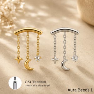 May include: Two curved barbell body jewelry pieces, one gold-toned and one silver-toned, each featuring three dangling chains with a four-pointed star, a crescent moon, and a round clear gemstone. Text reads: G23 Titanium Internally threaded.