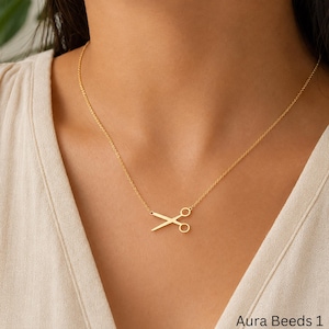 May include: A gold-toned necklace featuring a delicate chain and a pendant shaped like a pair of open scissors. The pendant is centered on the chain, which rests against the collarbone. The text Aura Beeds 1 is visible in the bottom corner.