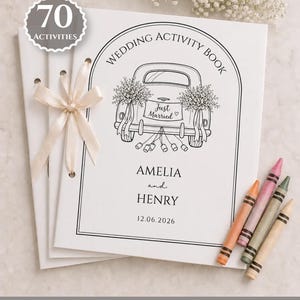 May include: A white wedding activity book with a black line drawing of a car with the text Just Married, Amelia and Henry, and 12.06.2026. The book is tied with a cream ribbon and includes four crayons in orange, pink, green, and tan colors.