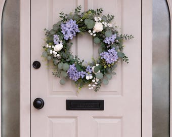May include: A floral wreath with purple, white, and green foliage hangs on a light gray door. The wreath features purple hydrangeas, white roses, and eucalyptus leaves. The door has a black handle and a mail slot that reads "LETTERS".