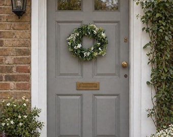May include: A gray front door with a wreath of greenery and white flowers. The door has a brass mail slot and a doorknob. A black lantern hangs to the left, and a small welcome mat sits on the doorstep. Plants frame the door.