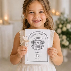 May include: A white rectangular wedding activity book with a black line drawing of a car with Just Married on the license plate. The cover features the text Wedding Activity Book, Amelia and Henry, and the date 12.06.2026, tied with a cream ribbon.