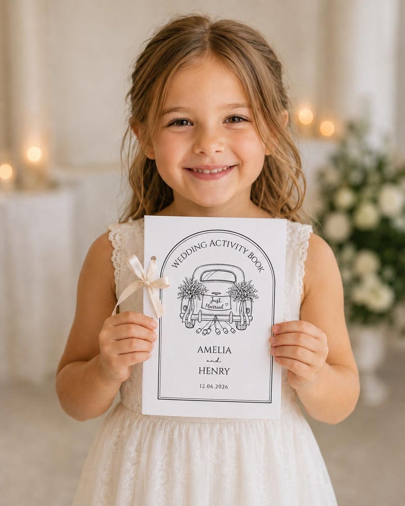 May include: A white rectangular wedding activity book with a black line drawing of a car with Just Married on the license plate. The cover features the text Wedding Activity Book, Amelia and Henry, and the date 12.06.2026, tied with a cream ribbon.