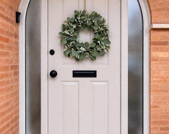 May include: A white front door with a decorative wreath made of green leaves and small white flowers. The door has a black handle and a mail slot. A black lantern hangs above the door.