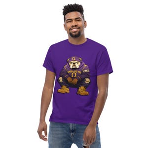 Puede incluir: Una camiseta morada con un bulldog de dibujos animados que lleva una sudadera con capucha morada con el texto "OMEGA PSI PHI" y la letra griega Omega. El bulldog lleva botas marrones y está en cuclillas. La camiseta la lleva una persona.