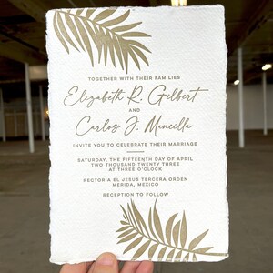 May include: A white wedding invitation with gold foil lettering and palm leaf designs. The invitation announces the marriage of Elizabeth R. Gilbert and Carlos J. Mencilla on Saturday, April 15, 2023, at 3:00 PM at the Rectoría El Jesús Tercera Orden in Mérida, Mexico. The invitation includes the text "Reception to follow."