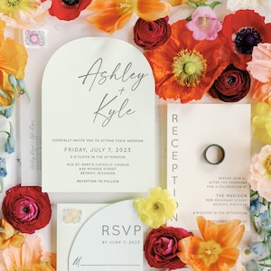 May include: Wedding invitation suite with a floral theme. The invitation features a white arch with the names "Ashley + Kyle" in gray script. The reception card features the text "Reception" in vertical black letters and the text "The Madison" in black. The RSVP card features the text "RSVP by June 7, 2023" in black.