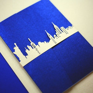 May include: A blue card with a white paper cutout of the New York City skyline. The skyline is attached to the card with a white ribbon.