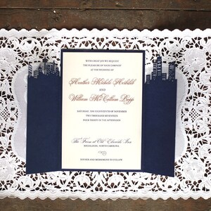 Puede incluir: Una invitaci&oacute;n de boda azul marino con una superposici&oacute;n de encaje blanco. La invitaci&oacute;n presenta una silueta de ciudad y el texto "With great joy we request the pleasure of your company at the wedding of Heather Michele Hershbell and William McCalium Pope Saturday, the eighteenth of November Two thousand seventeen Four thirty in the afternoon The Farm at Old Elmore Inn Highlands, North Carolina Dinner and Merriment to Follow"