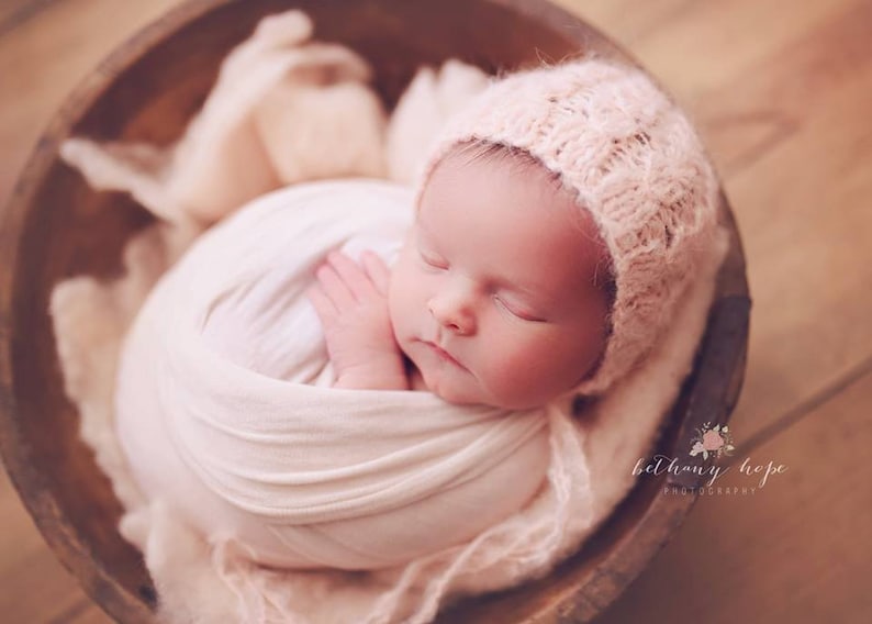 May include: A newborn baby sleeping peacefully in a wooden bowl, wrapped in a soft white blanket and wearing a crocheted bonnet. The baby's face is visible, with closed eyes and a peaceful expression.