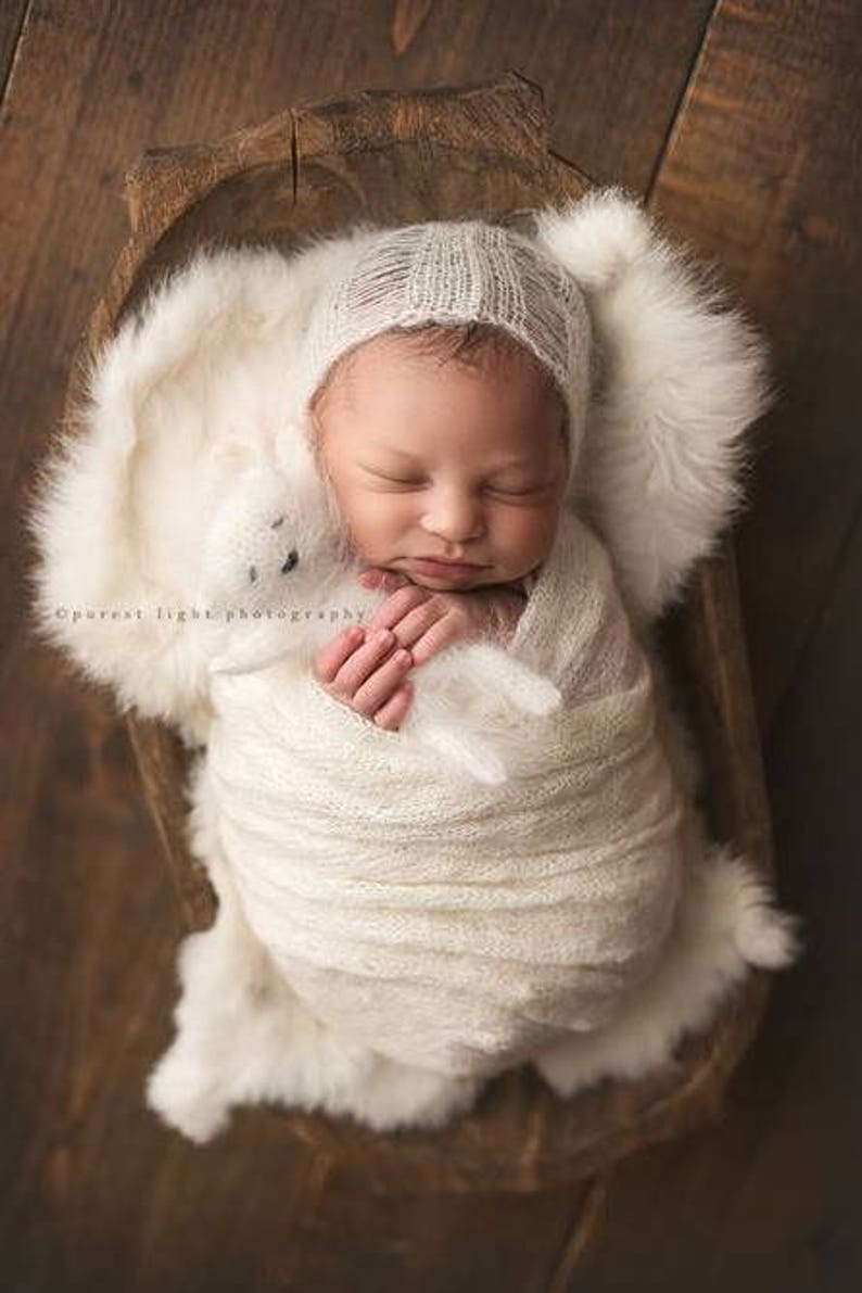 May include: A newborn baby is swaddled in a white knitted blanket and wearing a white knitted hat. The baby is sleeping peacefully in a wooden bowl lined with white faux fur. The baby is holding a white knitted toy.