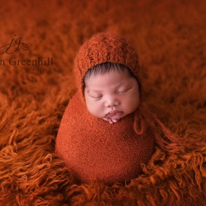 May include: A newborn baby is swaddled in a rust-colored blanket and wearing a matching knitted hat. The baby is sleeping peacefully on a fluffy orange rug.