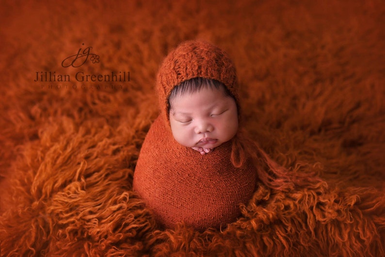 May include: A newborn baby is swaddled in a rust-colored blanket and wearing a matching knitted hat. The baby is sleeping peacefully on a fluffy orange rug.