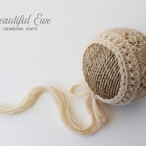 Könnte beinhalten: Eine beige gestrickte Babyhaube mit einem gehäkelten Rand und einer langen, cremefarbenen Garnbinde. Die Haube ist aus einem strukturierten Garn gefertigt und hat ein zartes, spitzenartiges Design. Der Text "Beautiful Eve Newborn Knits" ist in der oberen linken Ecke des Bildes sichtbar.