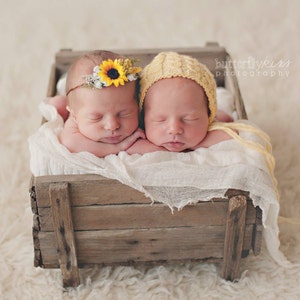 May include: Two newborn babies sleeping in a wooden crate. One baby is wearing a yellow crocheted hat and the other is wearing a sunflower headband.