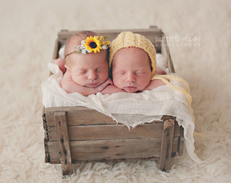 May include: Two newborn babies sleeping in a wooden crate. One baby is wearing a yellow crocheted hat and the other is wearing a sunflower headband.