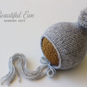 Puede incluir: Un gorro de bebé de punto gris con un pompón en la parte superior y una larga borla gris. El gorro está hecho por Beautiful Ewe Newborn Knits.