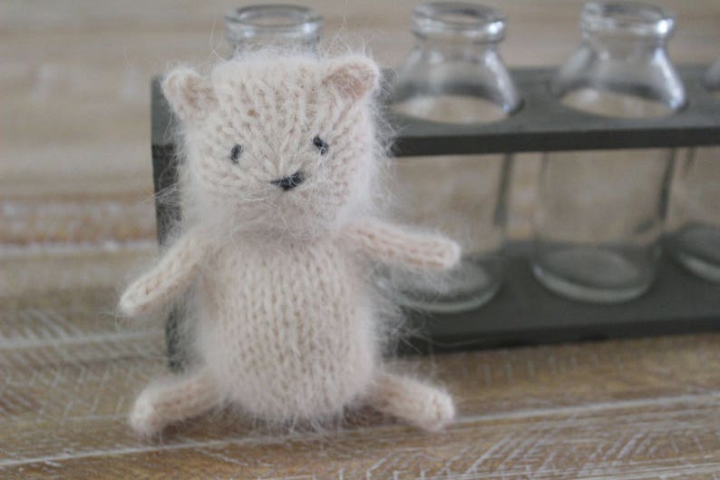 May include: A soft, white knitted toy animal with black eyes and a black nose. The toy is sitting on a wooden surface in front of a metal rack with glass bottles.