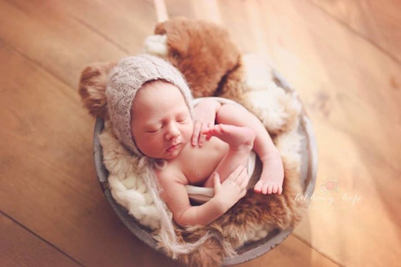 May include: A newborn baby wearing a knitted hat sleeps peacefully in a basket lined with faux fur. The baby is curled up with its legs tucked in and its arms wrapped around itself.