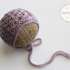 May include: A purple knitted baby bonnet with a lace pattern and a long tie. The bonnet is displayed on a woven straw ball.